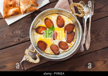 Türkisches Frühstück - Spiegeleier mit Würstchen (sucuk) und Gewürzen in einer Pfanne auf einem hölzernen Hintergrund. Stockfoto