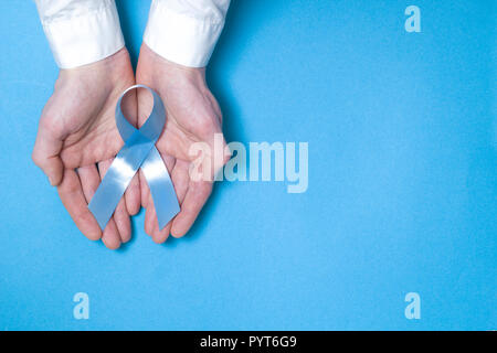 Das Symbol der Männer, die von Prostatakrebs betroffen ist ein Light Blue Ribbon. Ein Tape in die Hände von einem Mann auf einem blauen Hintergrund. Kopieren Sie Platz. Stockfoto