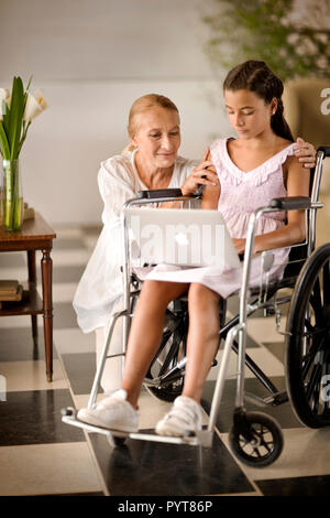 Junge Mädchen im Rollstuhl, etwas Übersicht zu ihrer Großmutter auf einem Laptop. Stockfoto