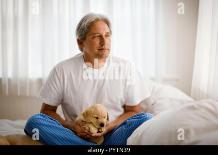 Entspannt im mittleren Alter Mann mit Labrador Welpe sitzend auf seinem Bett. Stockfoto