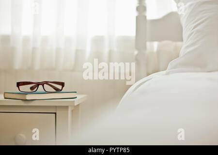 Buch und Brille auf einem Nachttisch neben dem Bett in einem Schlafzimmer. Stockfoto