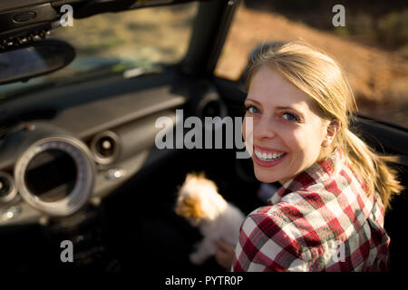 Porträt eines lächelnden jungen Frau auf dem Beifahrersitz ein Cabrio Auto sitzen mit ihrem kleinen Hund. Stockfoto