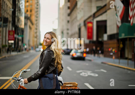 Portrait von eine fröhliche junge Frau, die ihr Fahrrad entlang einer belebten Straße der Stadt. Stockfoto