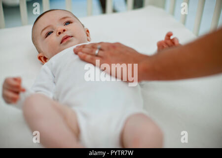 Cute Baby Boy schauen aus seinem Kinderbett als seine junge Mutter Stellen sanft eine Hand auf seine Brust. Stockfoto