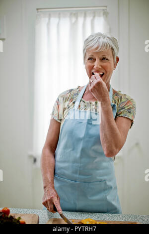 Heiter, reife Frau genießt Snacking auf ein Stück frisches Gemüse wie Sie bereitet ein gesundes Mittagessen. Stockfoto