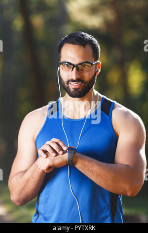 Junge athletische multikulturellen bärtigen Mann mit sportish Gläser, die Pause nach der morgendlichen Joggen, er Musik hören und schauen auf seine sportish beobachten, gesunden Lebensstil und Personen Konzept Stockfoto