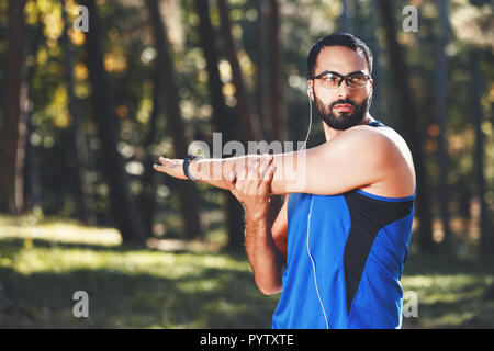 Junge athletische multikulturellen bärtigen Mann mit Brille sportish seine Lieblingsmusik hören, während seine Arme Stretching, gesunde Lebensweise und Personen Konzept Stockfoto