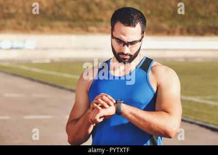 Athletische multikulturellen bärtigen Mann mit sportish Gläser auf die Indikatoren auf seinem sportish Watch auf dem Stadion, gesunden Lebensstil und Menschen Konzept Stockfoto