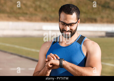 Athletische multikulturellen bärtigen Mann mit Brille sportish lächelnd, während sie auf die Indikatoren auf seinem sportish Watch auf dem Stadion, gesunden Lebensstil und Menschen Konzept Stockfoto