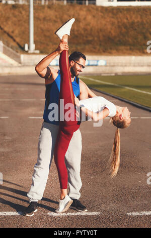 Multikulturelle gut aussehender bärtiger Mann im blauen Hemd Hilfe Stretching zu seinen blonden athletische Freundin am Fußball Stadium im Freien, gesunden Lebensstil und Menschen Konzept Stockfoto