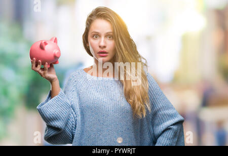 Junge hübsche blonde Frau mit Sparschwein über isolierte Hintergrund im Schlag mit einer Überraschung konfrontiert, ängstlich und aufgeregt mit Angst Angst Ausdruck Stockfoto