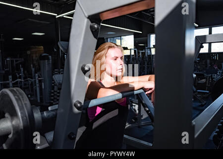 Passen junge Frau, die an der Turnhalle vor Barbell. Weibliche Athlet Porträt im Fitnessraum unter viele Fitnessgeräte Stockfoto