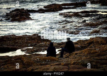 Ein älteres Paar sitzen in der Nähe des Ozeans, die Felsen und hohe Wellen. Ansicht von hinten. Stockfoto