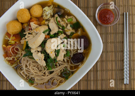 Chinesische Nudelsuppe Gericht in Quadratische weiße Schüssel. Kleines Glas Schale mit pikanten Soße und Essstäbchen auf Holz- mat. Stockfoto