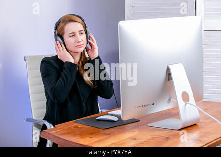 Porträt der glückliche junge erfolgreiche Geschäftsfrau im Büro. Sie sitzt am Tisch mit Kopfhörern und mit Blick auf die Anzeige. Musik hören Stockfoto