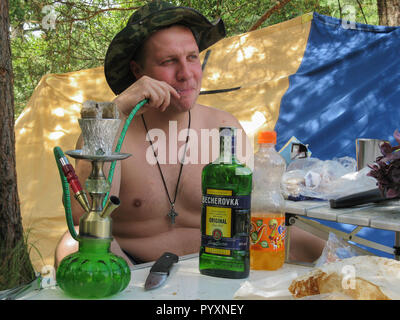 Ein Mann in einem Hut raucht eine Shisha und Alkohol trinkt. Camping. Stockfoto