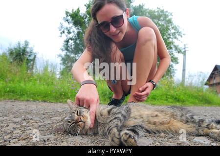 Junge Frau in Sonnenbrille petting Grau gestreifte Katze auf der Straße. Stockfoto