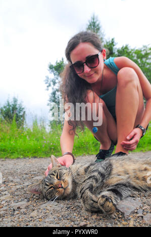 Junge Frau in Sonnenbrille petting Grau gestreifte Katze auf der Straße. Stockfoto