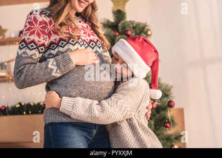 Zugeschnittenes Bild der Tochter in santa hut Umarmen schwangeren Mutter in der Nähe von Weihnachten zu Hause Stockfoto