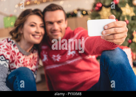 Selektiver Fokus der lächelnde Paar in Pullover unter selfie mit Smartphone in der Nähe von Weihnachten zu Hause Stockfoto