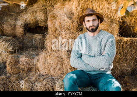 Zuversichtlich männlichen Landwirt in Pullover und Strohhut sitzt mit verschränkten Armen auf Heu Stapel an der Ranch Stockfoto