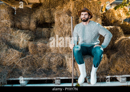 Nachdenklicher mann Landwirt in Pullover und Strohhut sitzend auf Heu Stapel an der Ranch Stockfoto