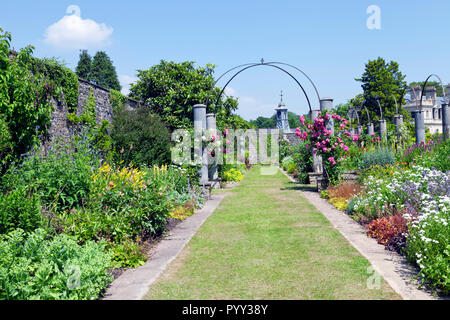 Bunte Blumen blühen auf beiden Seiten des Grases weg, Rosa unter einem Bogen, Bäume und Sträucher von einer alten Steinmauer, in üppigem Englischer Garten, auf einer s Stockfoto