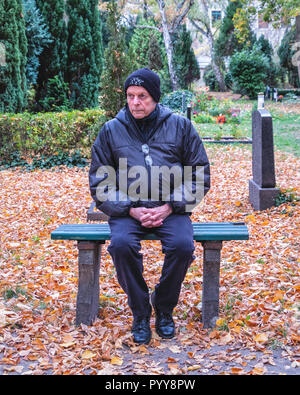 Älterer Mann sitzt auf dekorativen schmiedeeisernen & Holz Werkbank, im Friedhof - Besinnlich, nachdenklich, traurig. Stockfoto