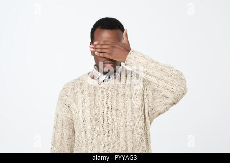 Afrikanischer Mann in Pullover bedeckte sein Gesicht mit den Händen über grauen Hintergrund. Stockfoto