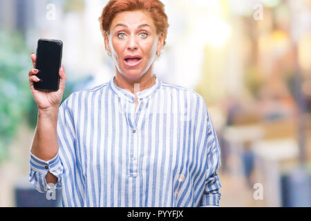 Attraktives senior kaukasischen rothaarige Frau, der Bildschirm des Smartphones über isolierte Hintergrund im Schock Angst mit einer Überraschung konfrontiert, ängstlich und aufgeregt Stockfoto