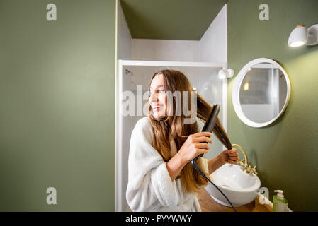 Junge und glückliche Frau im Bademantel Haarglättung mit Haarglätter im Badezimmer Stockfoto