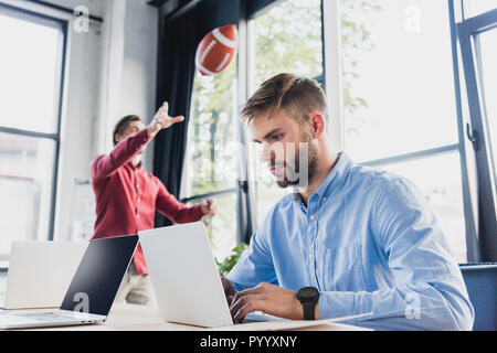 Junge Unternehmer mit Laptop, während Kollege spielen mit Rugby Ball hinter im Büro ausgerichtet Stockfoto