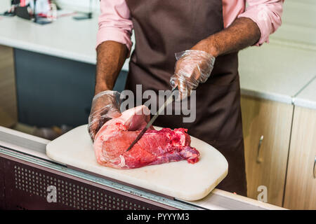 Zugeschnittenes Bild der afrikanischen amerikanischen männlichen Metzger in Schürze Schneiden von rohem Fleisch im Supermarkt Stockfoto