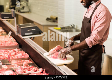 Teilweise mit Blick auf die lächelnde afrikanischer amerikanischer Mann Shop Assistant im Vorfeld steak Schneiden von rohem Fleisch im SB-Warenhaus. Stockfoto