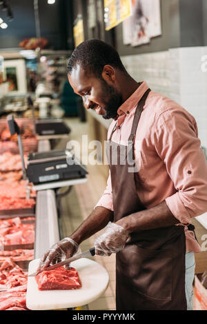 Lächelnd afrikanischer amerikanischer Mann Shop Assistant im Vorfeld steak Schneiden von rohem Fleisch im Supermarkt Stockfoto