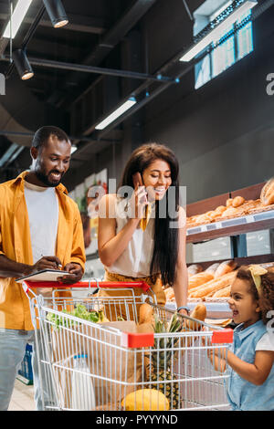 Lächelnd afrikanische amerikanische Frau im Gespräch auf dem Smartphone während ihrem Mann und ihrer Tochter in der Nähe von mit Einkaufswagen im Supermarkt Stockfoto