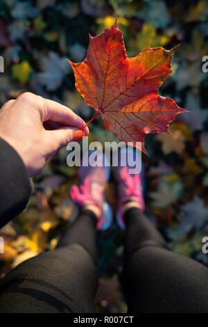 Frauen im Sport Kleidung Holding ein Blatt in der Hand. Nahaufnahme von Frau Ruhe und Entspannung im Herbst Park, sitzen auf den Boden unter gelbe Blätter. Konzept der Saison und Fallen. Stockfoto