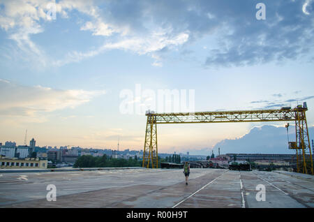 Unfinished Metal Bridge. Sportliche Mädchen mit Rucksack in Shorts und mit Dreadlocks. Industrielle Landschaft bei Sonnenuntergang verlassen Stockfoto