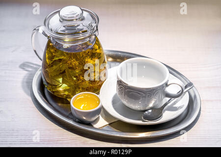 Hot linden - Blumen Tee in transparentem Glas Teekanne, kleine Tasse Honig, und leeren weißen Porzellan Schale und silbernen Löffel auf Untertasse auf Silber tra angeordnet Stockfoto