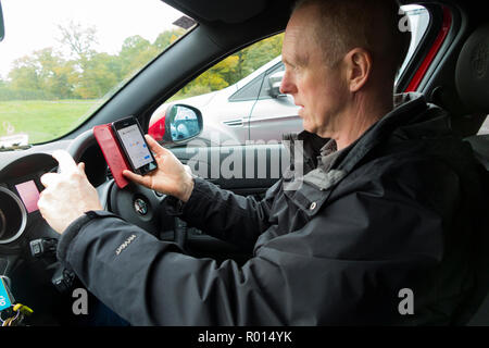 Im mittleren Alter Fahrer während der Fahrt telefonieren und mit einem mobilen Telefon Telefon Telefon/Internet, e-Mail/E-Mail, Surfen im Internet surfen ist eine illegale Handlung und könnte zu Punkten auf Lizenz oder Tod und Schädigung anderer Verkehrsteilnehmer führen. Großbritannien (103) Stockfoto