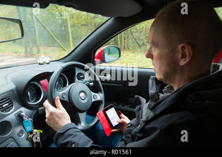 Im mittleren Alter Fahrer während der Fahrt telefonieren und mit einem mobilen Telefon Hörer eine SMS-Nachricht, die eine illegale Handlung zu senden und könnte zu Punkten auf Lizenz, oder Tod und Verletzungen von Fußgängern und anderen Verkehrsteilnehmern führen. Großbritannien (103) Stockfoto