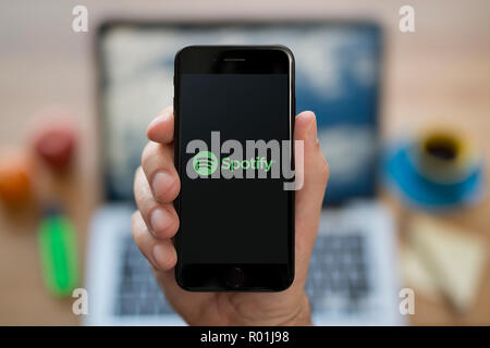 Ein Mann schaut auf seinem iPhone die zeigt die Spotify logo, während saß an seinem Computer Schreibtisch (nur redaktionelle Nutzung). Stockfoto