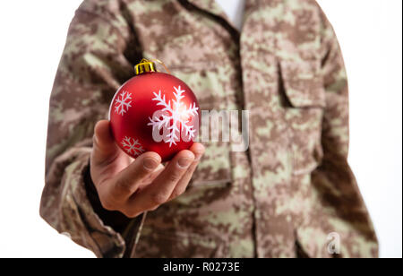 Weihnachten und Armee. Junge Soldat hält ein rotes christmas Ball stehend auf weißem Hintergrund Stockfoto