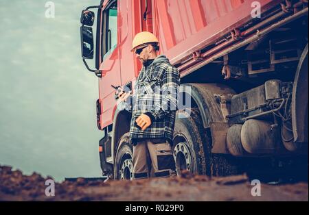 Kaukasische Dump Truck Fahrer in seinem 30s und die Baustelle voller Schmutz zu bewegen. Stockfoto