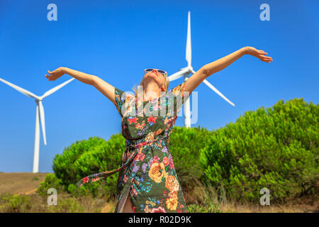 Eine blonde Frau mit offenen Armen mit Windkraftanlagen in Sagres, Algarve, Portugal, zu drehen. Alternative Energie, erneuerbare Energien und ökologische Nachhaltigkeit. Stockfoto