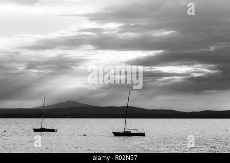 Ein paar leer, kleinen Segelboot auf einem See, unter einem Moody Himmel mit Sonnenstrahlen durch Filterung. Stockfoto