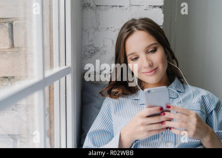 Lächelnd attraktive Frau kreiert neue Wiedergabeliste auf dem Mobiltelefon, E-Mail und genießt die Musik im Kopfhörer, sitzt auf der Fensterbank in einem der gemütlichen Zimmer, genießt Loni Stockfoto