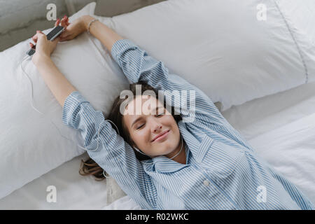 Gerne junge europäische Frau erstreckt sich im Bett, mit geschlossenen Augen, in gestreiften nightclothes Gekleidet, hält Smart Phone, genießt Lieblingsmusik in Ohrhörer Stockfoto