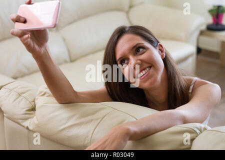 Hübsche brünette Mädchen, stützte sich auf ihren Arm lächelte ihr Mobiltelefon, mit dem Sie eine sehr helle und feminin selfie Foto suchen Stockfoto