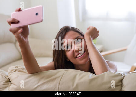 Hübsche brünette Mädchen, stützte sich auf ihren Arm lächelte ihr Mobiltelefon, mit dem Sie eine sehr helle und feminin selfie Foto suchen Stockfoto